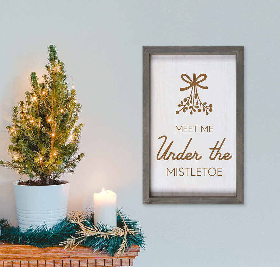 bordered wood sign that says meet me under the mistletoe mounted to the wall next to a small lit tree and a candle sitting on the mantle