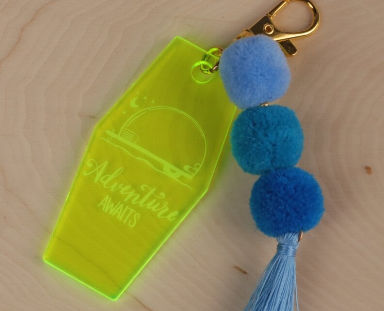 Bright keychain engraved with Adventure Awaits and a tassel of three blue pom-poms