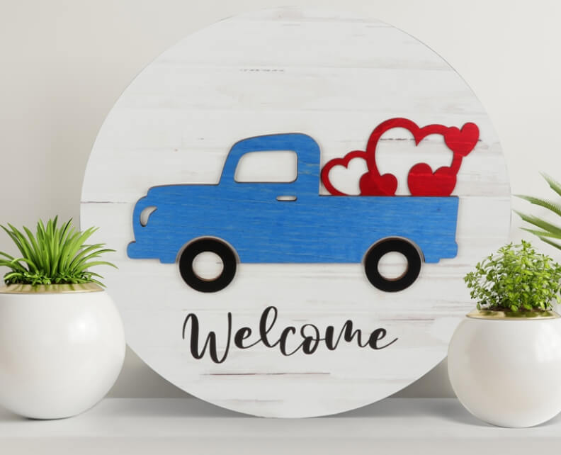 Wooden round home decor wall hanging with a pickup carrying hearts that says Welcome
