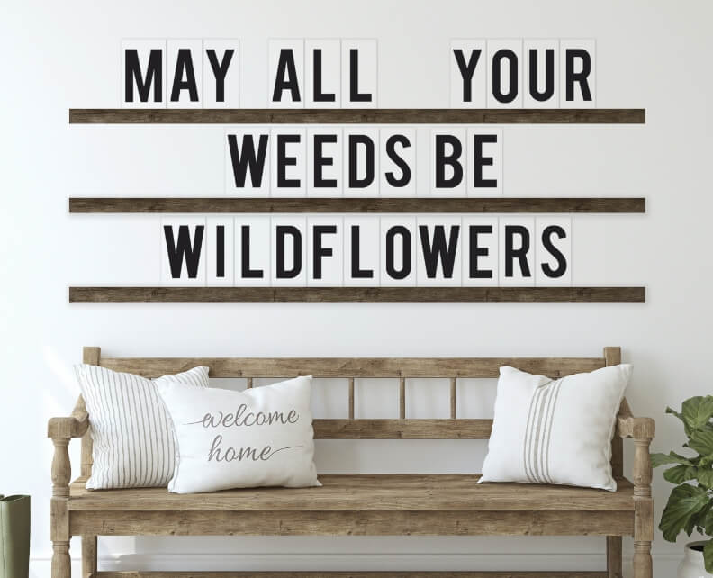 Wall decor, May all your weeds be wildflowers, above a wooden bench with cushions on it