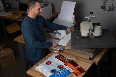 Man placing items in a heat press, standing next to a printer at a work table